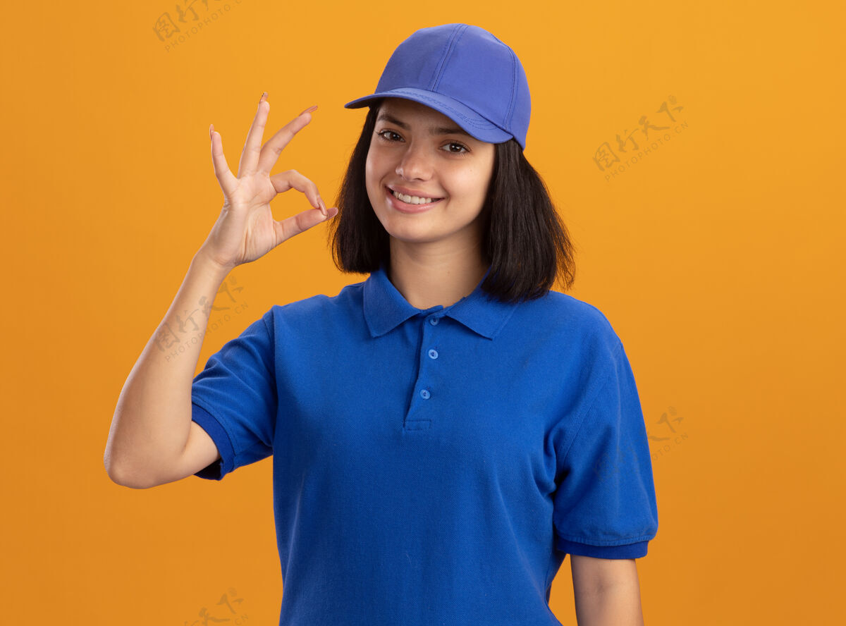 帽子身着蓝色制服 头戴鸭舌帽 站在橘色墙壁上微笑着表示同意的年轻送货女孩制服站着交货