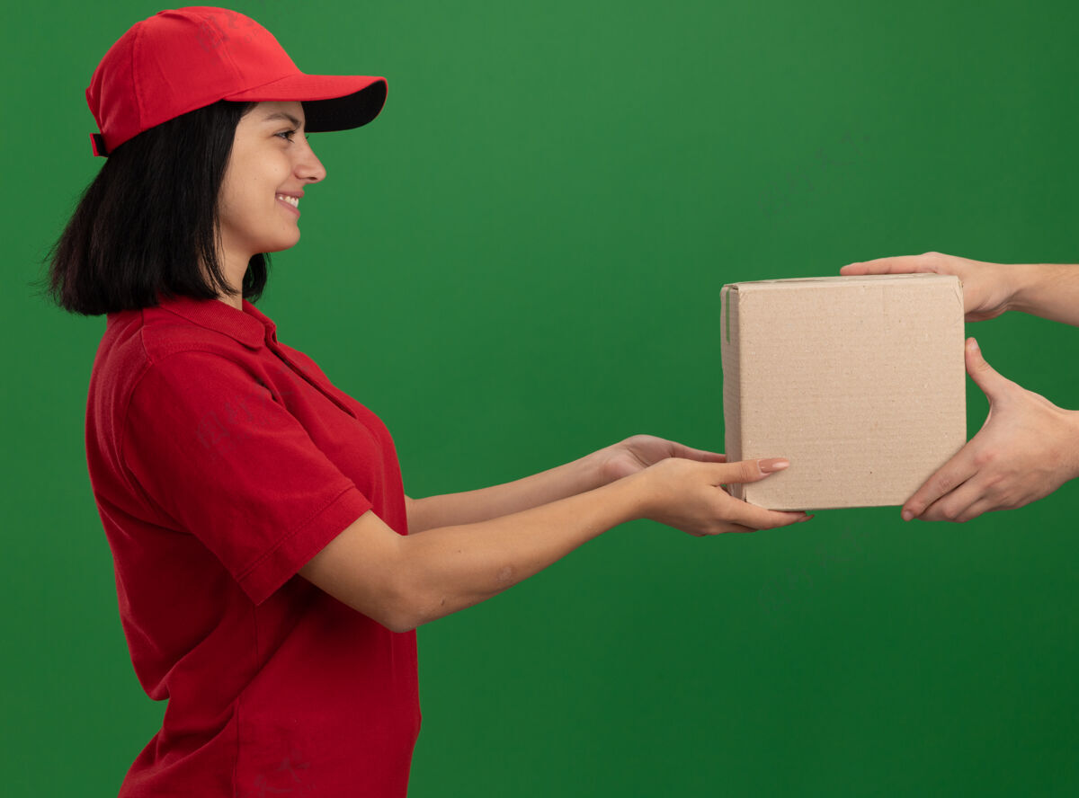 顾客身穿红色制服 头戴鸭舌帽的年轻送货员站在绿色的墙上 微笑着向顾客赠送纸板箱女孩制服给予