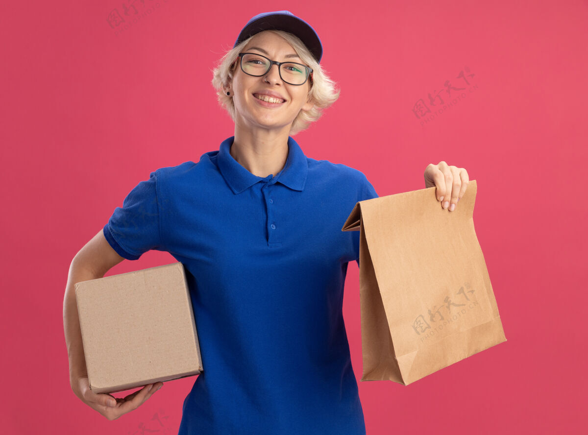包装穿着蓝色制服 戴着帽子 手里拿着纸包和纸板箱的快乐的年轻女送货员在粉红色的墙上欢快地笑着微笑帽子年轻