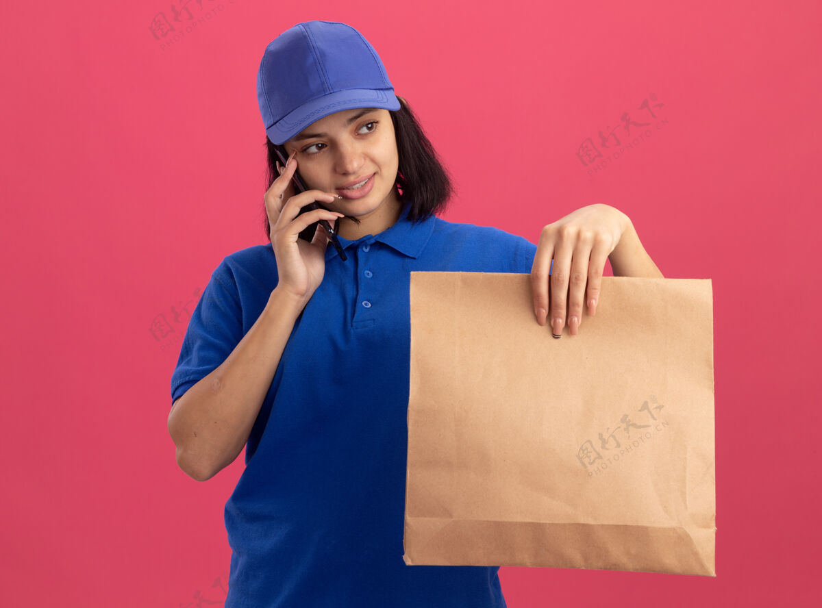 帽子身穿蓝色制服 戴着帽子 拿着纸包的年轻送货员站在粉红色的墙上 面带微笑地讲着手机微笑电话拿着