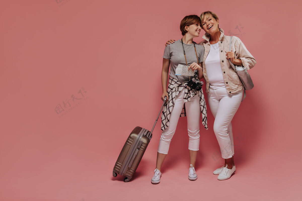白色迷人的两位女士 短而酷的发型 穿着轻薄的现代服装 拿着票 相机和手提箱摆姿势 在粉色背景上微笑着衬衫旅行成人