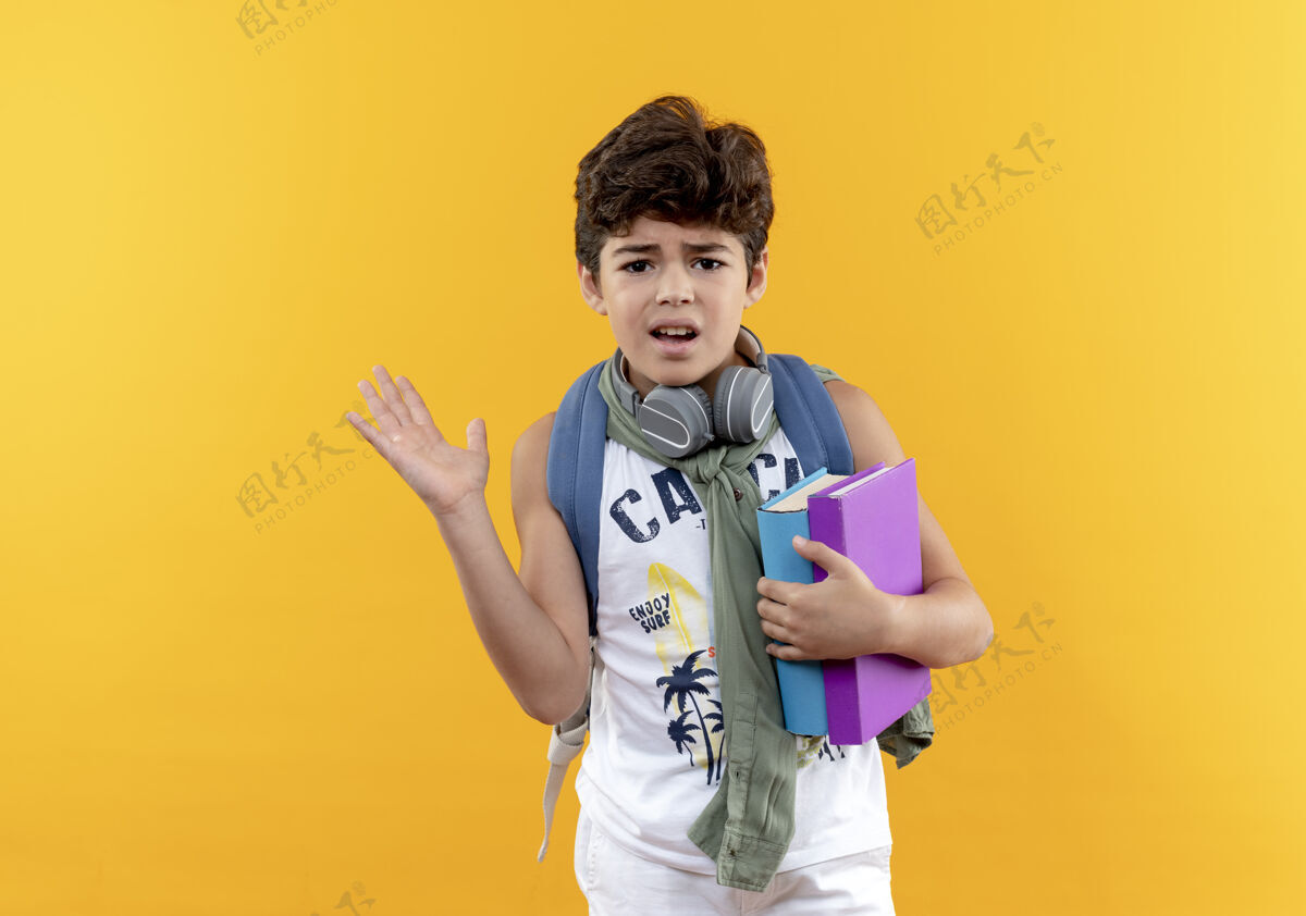 戴困惑的小男孩戴着书包和耳机拿着书 用手指着隔离在黄色背景上的一边 还有复印空间包学校耳机