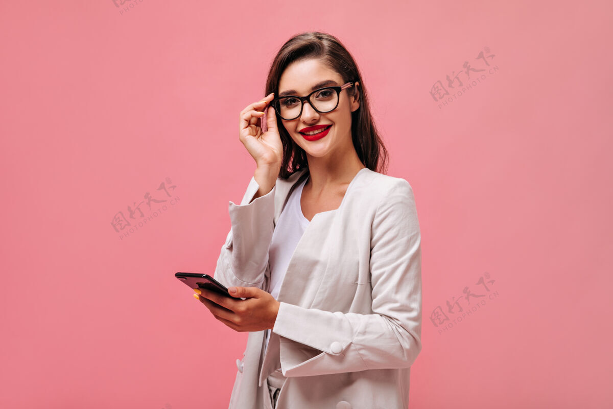 年轻迷人的年轻女子 红唇 米色套装 戴着眼镜 看着相机 拿着手机 背景是孤立的粉色休闲空白站立