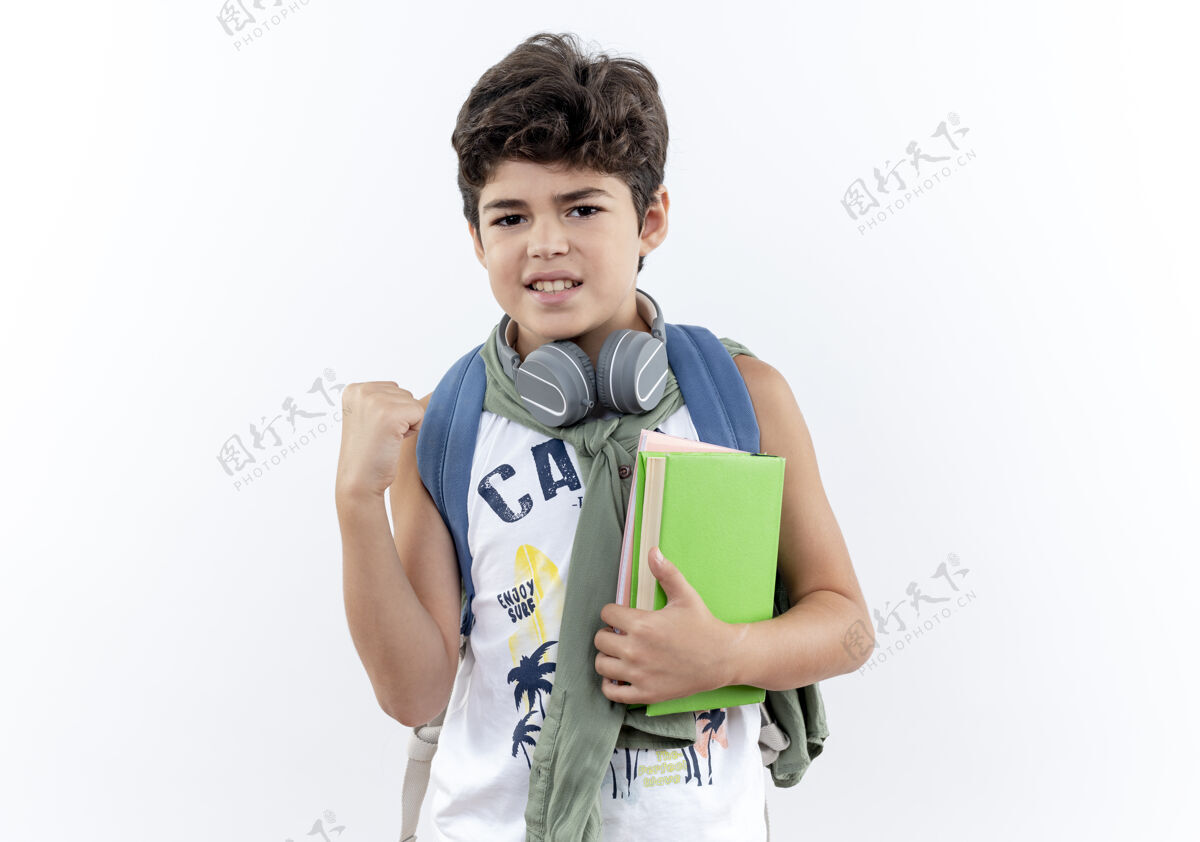 小学生看着镜头 一个背着书包 戴着耳机 手拿书本 手指交叉的小男孩被隔离在白色背景上小包手指