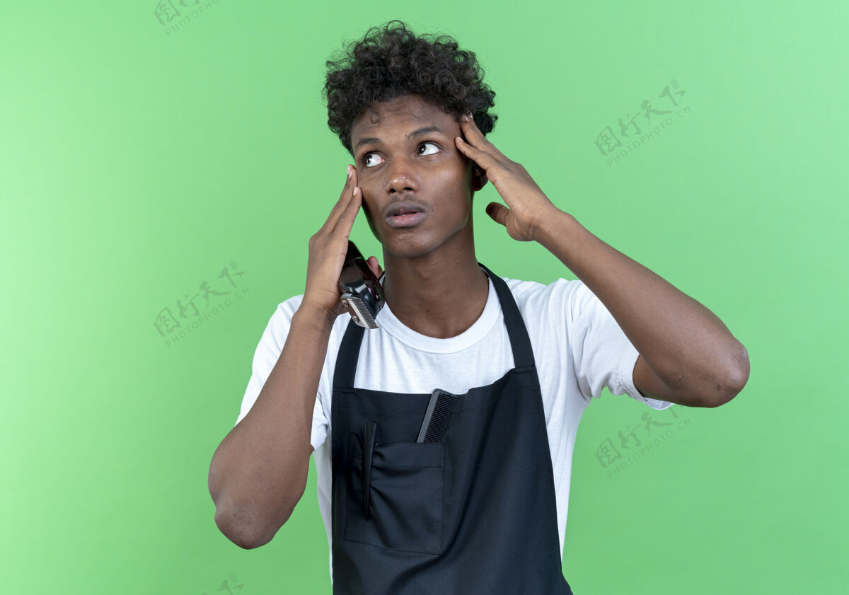 额头看着身边的年轻美籍黑人男理发师穿着制服拿着理发器 把手指放在额头上 隔离在绿色的背景上印象非裔美国人年轻