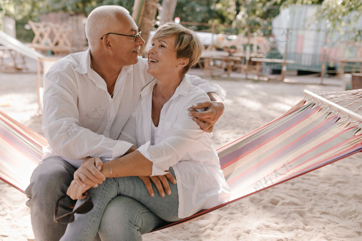 肖像快乐的老妇人 金发碧眼 穿着衬衫和牛仔裤 坐在格子吊床上 在沙滩上和微笑的白发男人拥抱祖父母海滩拥抱
