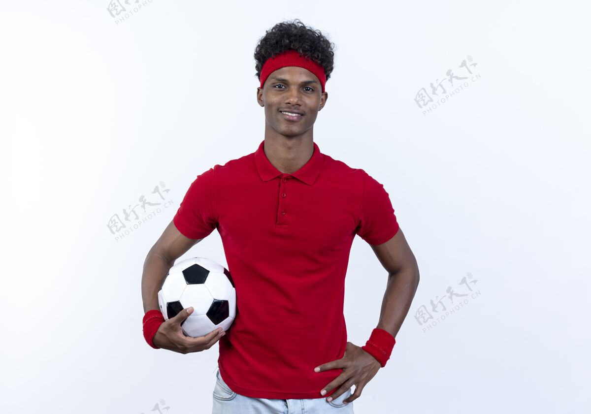 运动高兴的年轻黑人美国运动男子戴着头带和腕带拿着球 把手放在臀部隔离在白色背景上年轻非裔美国人臀部