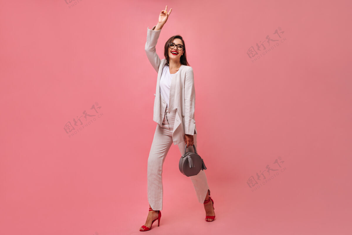 包一位身着白色现代西服 戴着眼镜 面带微笑的迷人女士手持手提包 在粉红色的背景上显示和平标志深色电话女性