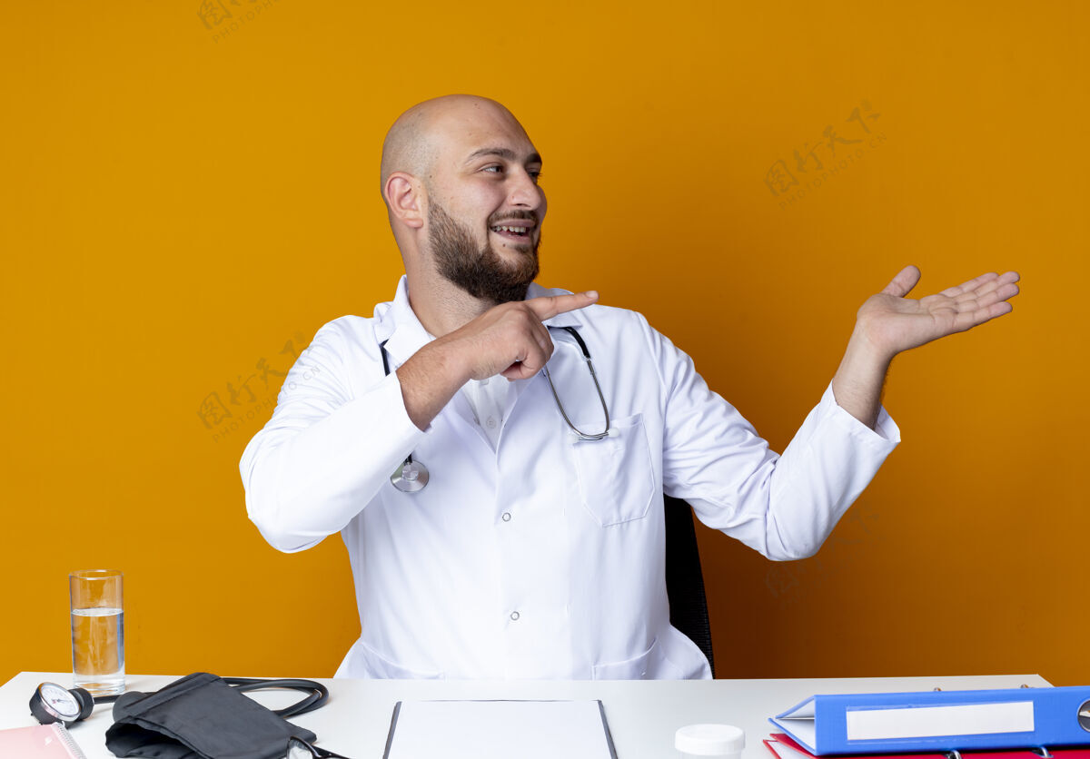工作高兴的年轻秃头男医生穿着医用长袍和听诊器坐在工作台上 手持医疗工具 假装拿着橙色背景上孤立的东西坐着工具办公桌