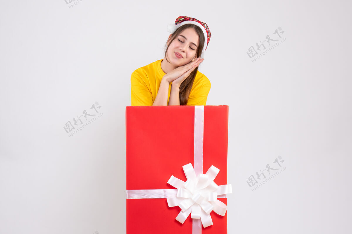 站前视图可爱的瞌睡女孩与圣诞老人帽子站在大礼物后面人节日圣诞老人