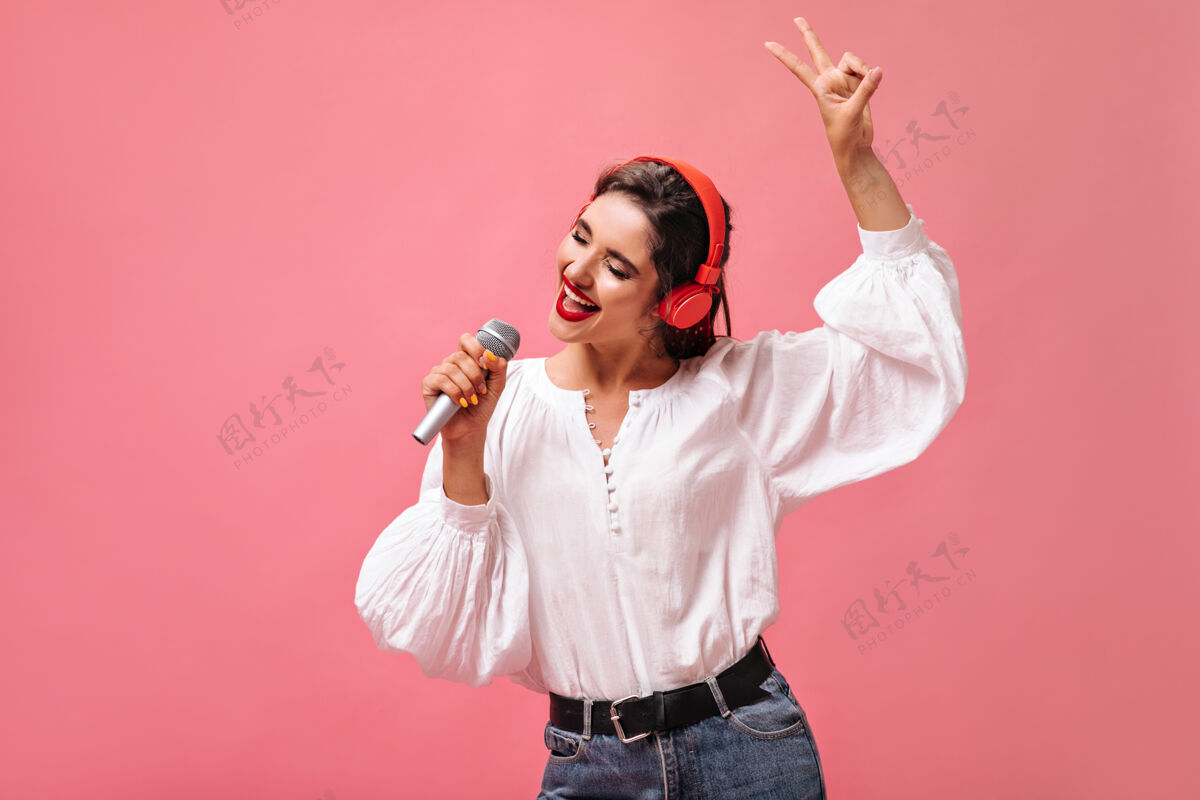 成人戴着红色耳机的漂亮女孩在粉色背景的麦克风里唱歌一位漂亮的女士 一头黑发 穿着时尚的白色衬衫 正在听音乐时尚肖像女性