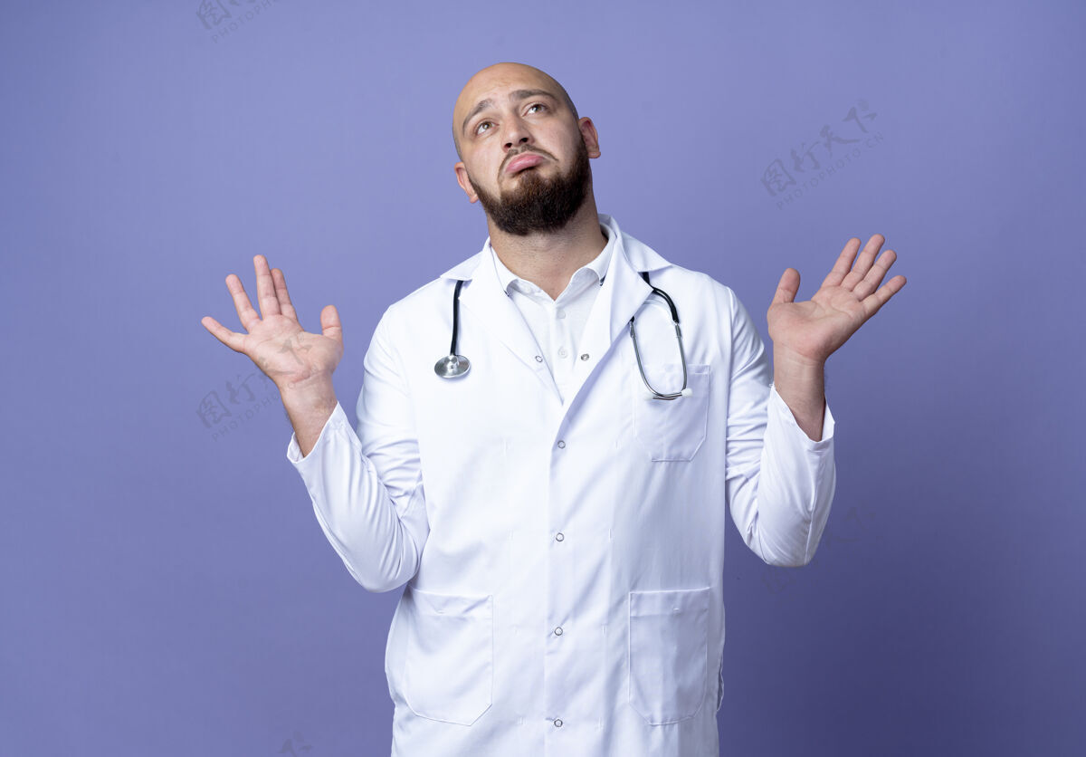 困惑抬头望着困惑的年轻秃头男医生 穿着医用长袍 听诊器摊开双手隔离在蓝色背景上传播穿听诊器