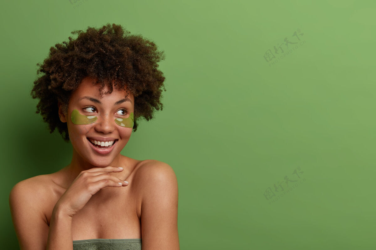 身体漂亮开朗的女人把手轻轻地放在下巴下 若有所思地看着旁边 戴着水凝胶眼罩 以保持健康新鲜的皮肤 裹着毛巾 隔离在绿色的墙上 复制文本空间美容贴片思想