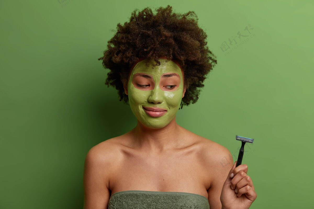 个人高兴的女人在家里涂保湿美容面膜 想拥有干净完美的皮肤 拿着剃须刀 做卫生处理 用毛巾裹着站在隔离的绿色墙壁上美容程序室内美容水疗