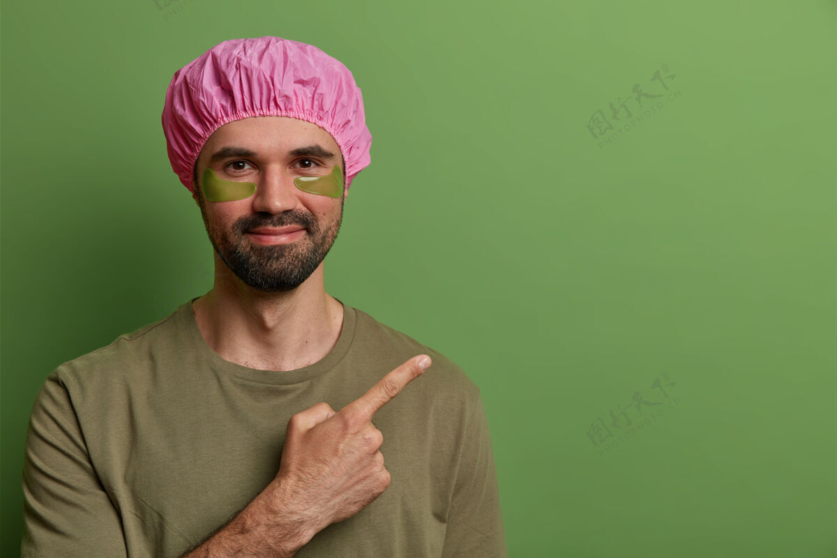 清洁满脸胡子的男人横观着粉色浴帽 休闲t恤 做眼部皮肤护理 用胶原蛋白垫减少细纹 指着空格 宣传一些产品面部帽子空白