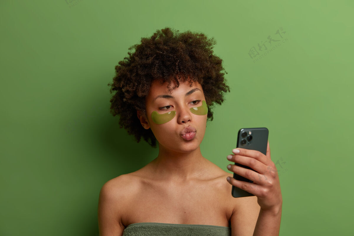 自拍年轻漂亮的女模特 卷曲的非洲头发 涂上水凝胶绿色贴片来减少黑眼圈 在手机上自拍 保持嘴唇圆润 用浴巾包着卫生非洲移动