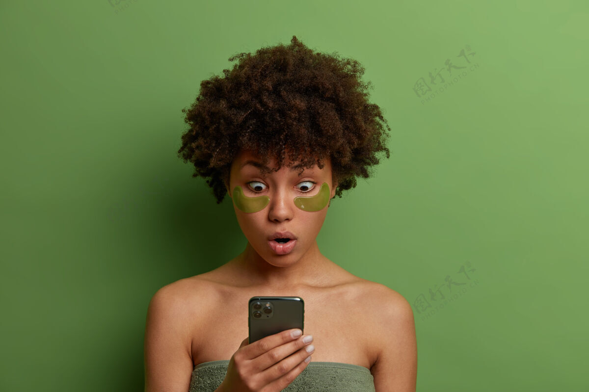 水凝胶美丽 护肤理念情绪激动的非裔美国妇女盯着智能手机屏幕 裹着毛巾站在那里 用眼罩来减轻浮肿 浏览互联网惊呆惊艳手机