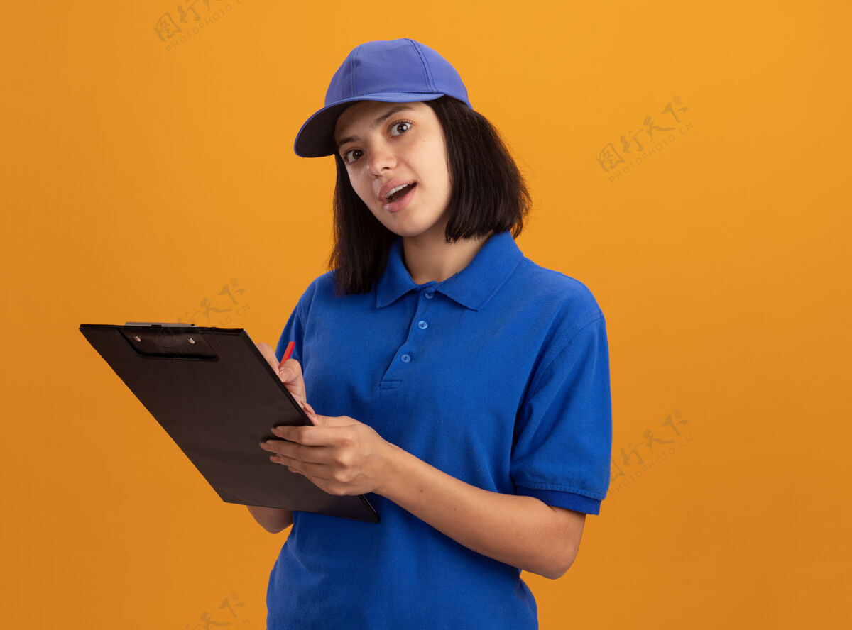 制服穿着蓝色制服 戴着帽子的年轻送货女孩站在橙色的墙上微笑着写着什么帽子站着写作