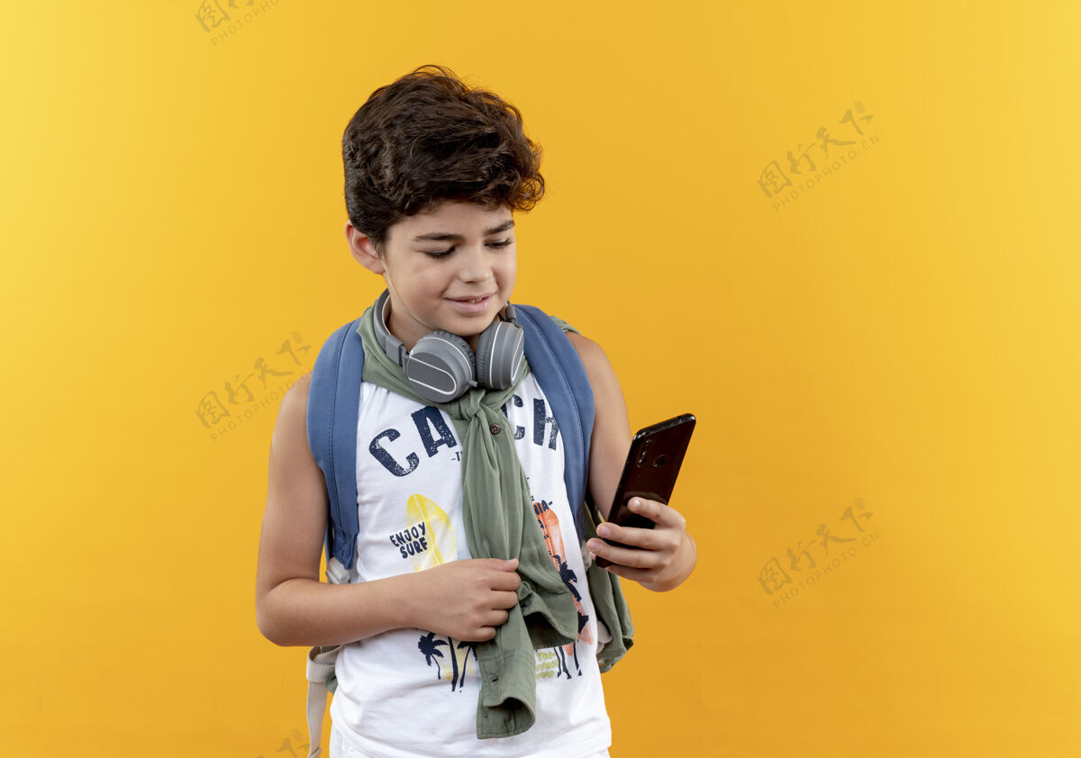 耳机高兴的小男生背着书包 戴着耳机 拿着手机看着黄色背景上的孤立男孩电话请