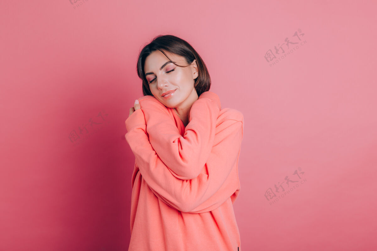 年轻笑容满面的年轻女子20多岁 穿着休闲服 在粉色背景的工作室里独立写真爱的概念闭着眼睛 双手交叉拥抱自己站立化妆拥抱