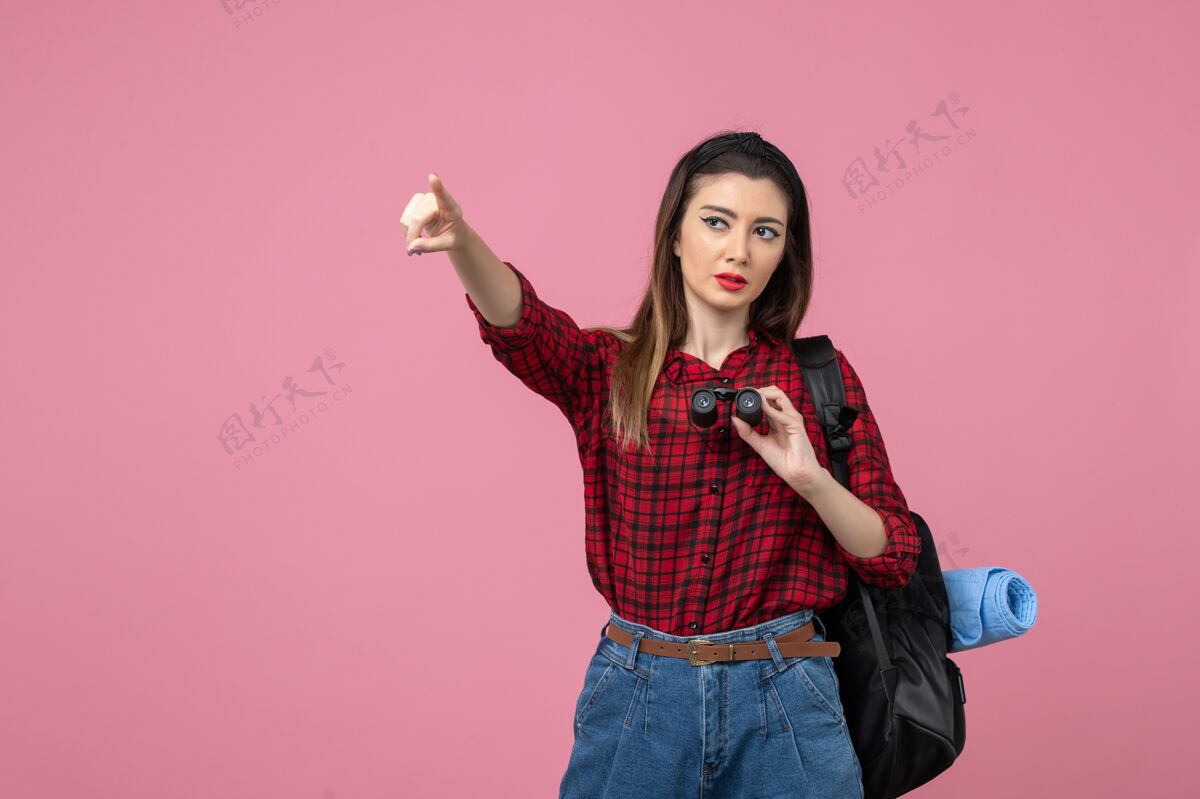 人正面图：穿着红色衬衫的年轻女性 用双筒望远镜在粉色背景上观察人类女性人年轻女性头发