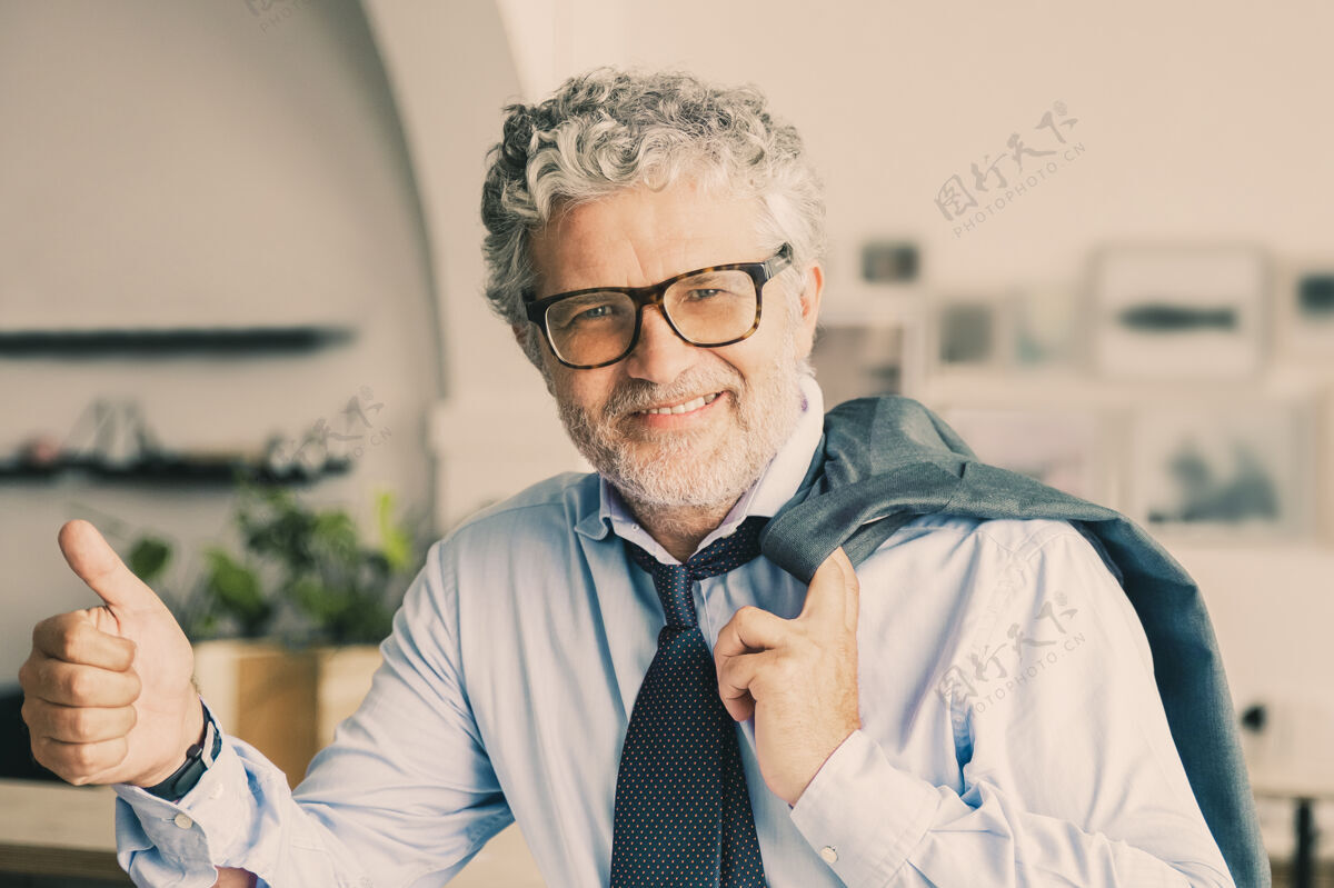 灰色快乐成熟的商务人士站在办公室咖啡厅 倚在柜台上 肩扛夹克 竖起大拇指男人商务人士竖起大拇指