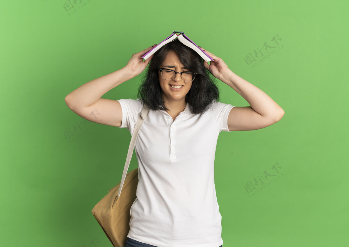 空间年轻气恼的漂亮白人女学生戴着眼镜 背着书包把书举过头顶放在绿色的空地上漂亮眼镜烦