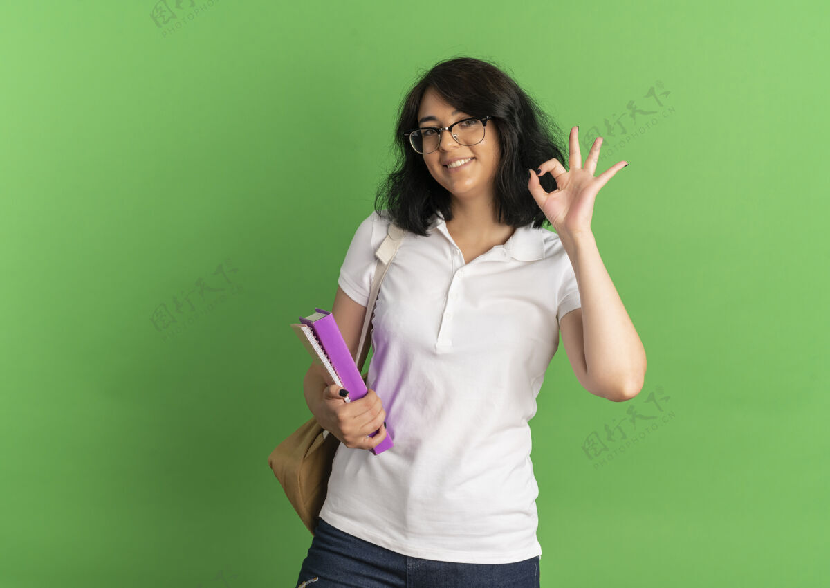 复制年轻的高加索女孩 面带微笑 戴着眼镜 背着书包 手势很好 手拿着书 在绿色的地方放着书年轻空间手势
