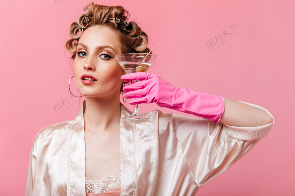 年轻一个穿着粉红色长袍的灰眼睛女人在隔离墙上拿着马提尼酒杯卷发时尚快乐