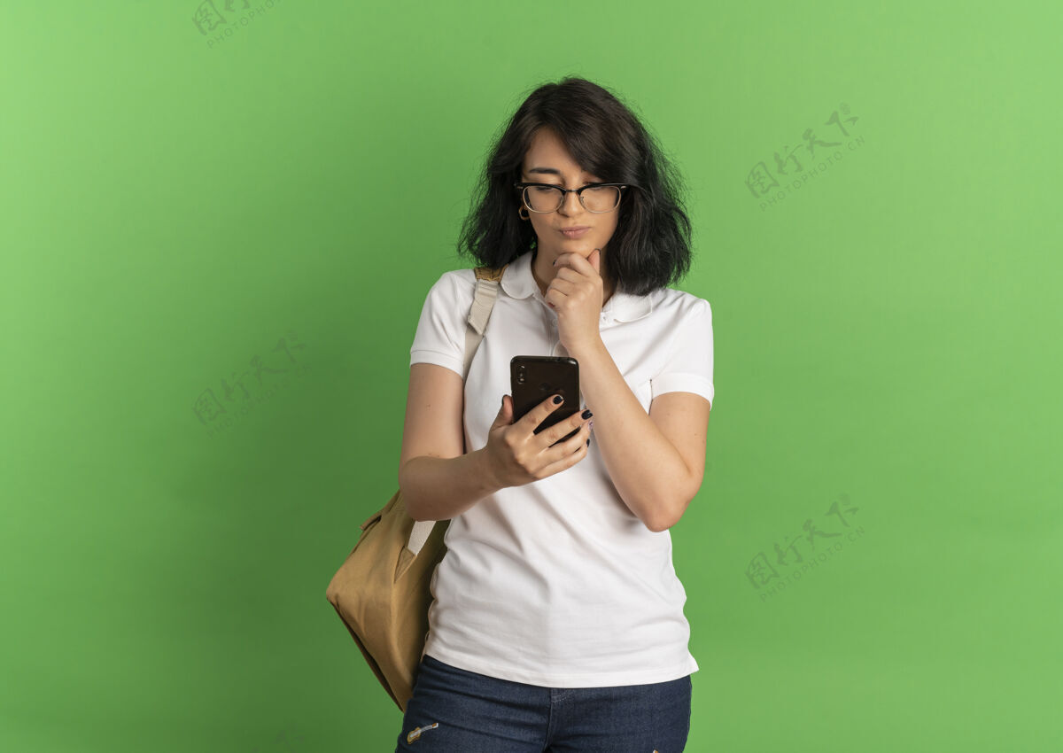 背部年轻体贴漂亮的白人女学生戴着眼镜 背着书包 手放在下巴上 看着绿色的手机和复印空间电话思想女学生