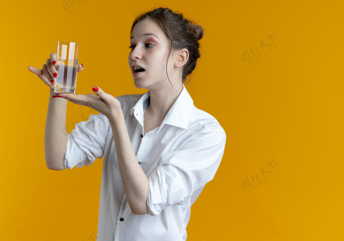 年轻年轻的惊讶的金发俄罗斯女孩拿着 指着一杯水在橙色的复制空间惊喜女孩空间