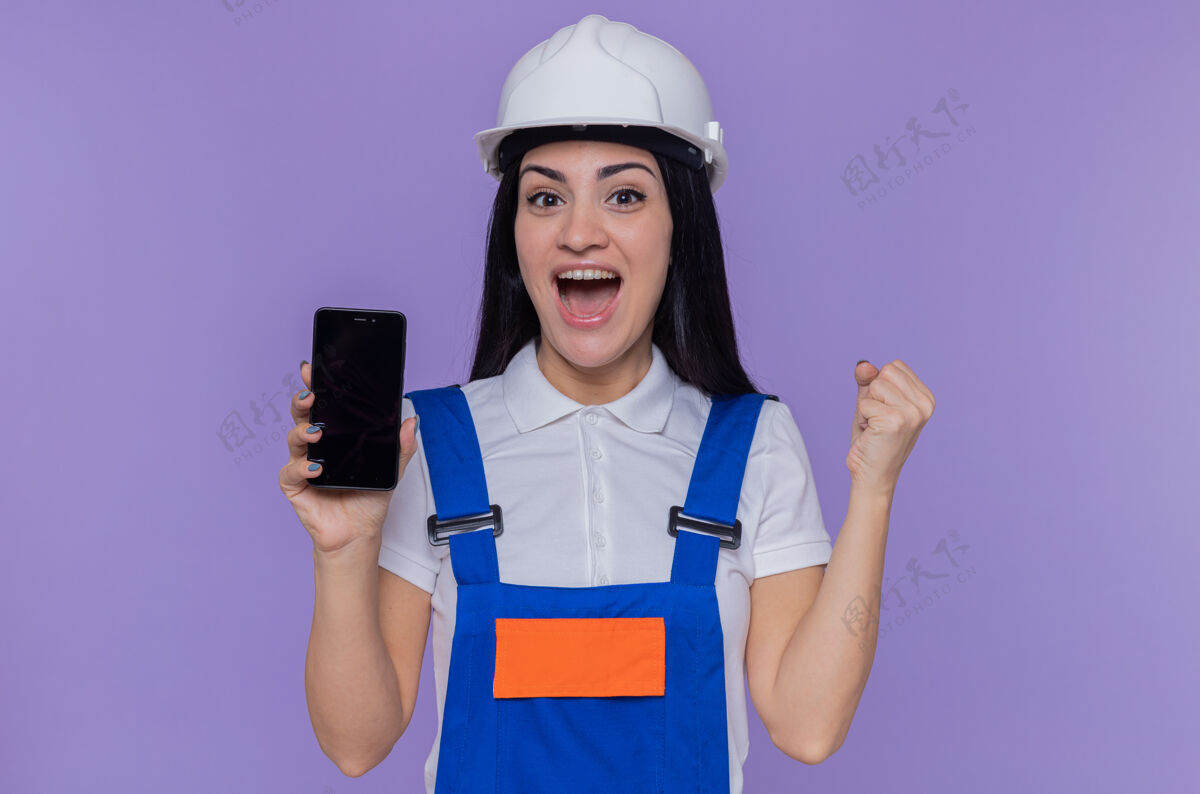 年轻身穿建筑制服 头戴安全帽的年轻建筑女工人正看着前方 高兴而兴奋地握紧拳头站在紫色的墙上移动制服兴奋