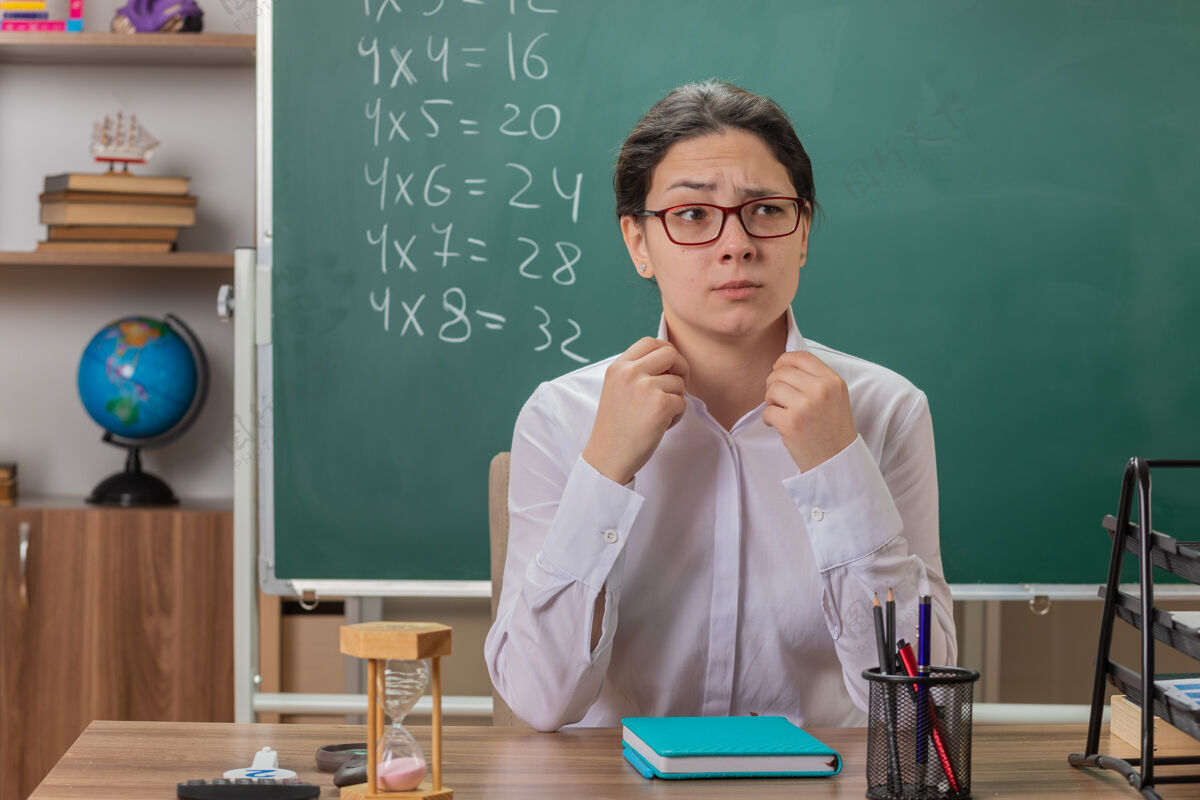 表情年轻的女老师戴着眼镜看着前面 带着自信的表情坐在教室黑板前的课桌上讲解课文学校戴着前面