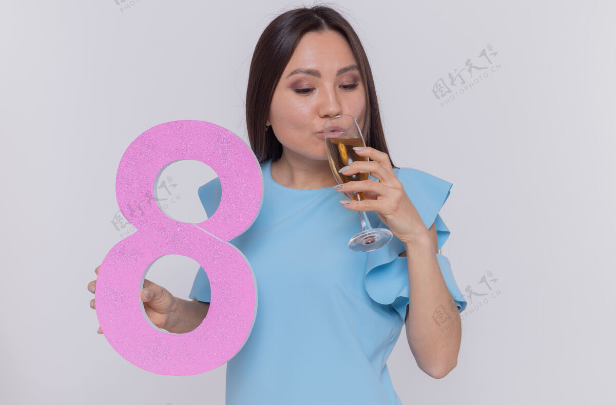 举行快乐的亚洲女人穿着蓝色的裙子 拿着八号硬纸板和香槟酒杯 站在白色的墙上庆祝国际妇女节杯子国际站