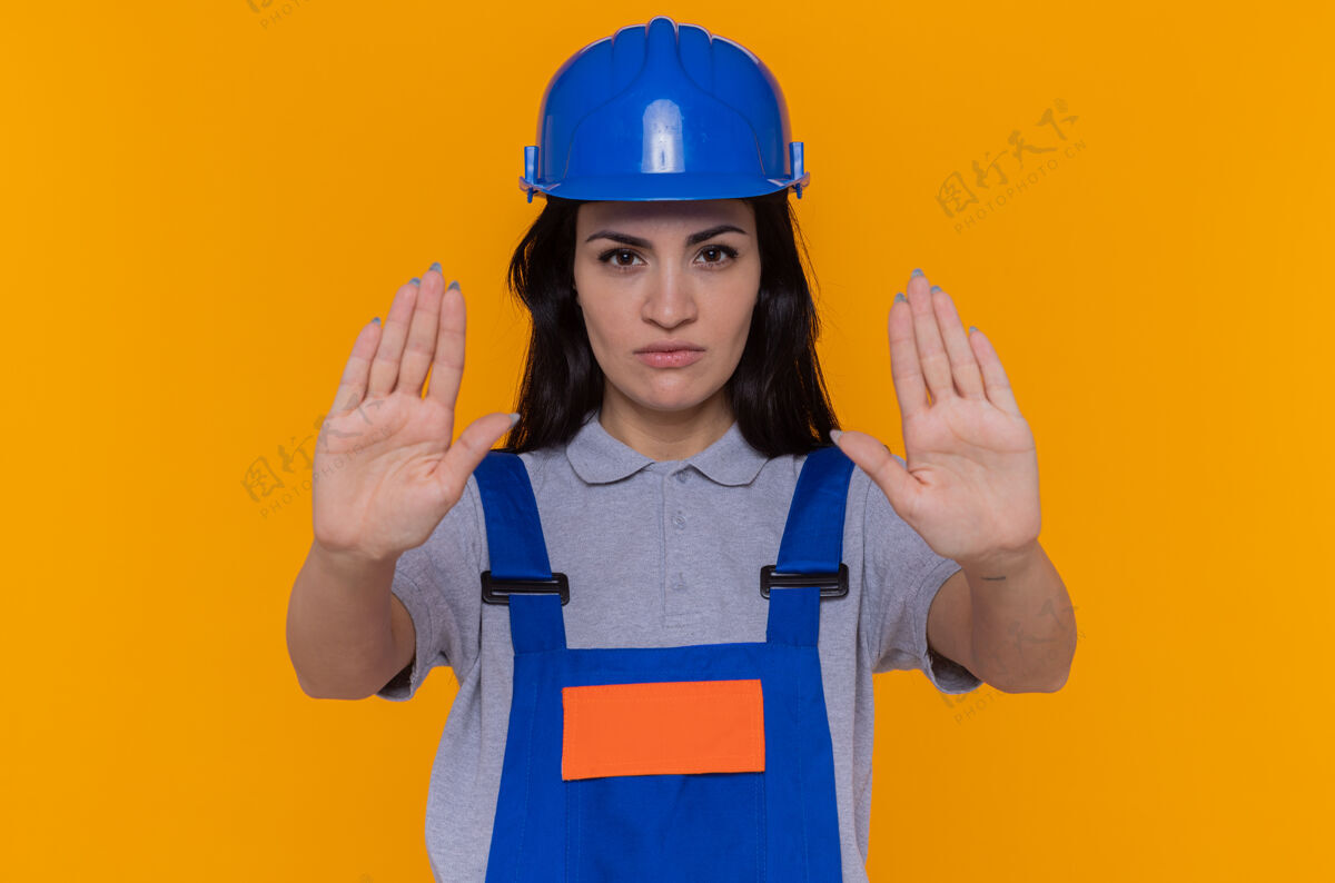 严肃身穿施工制服 头戴安全帽的年轻建筑女工人严肃地看着前方 双手站在橘色的墙上 做着停车的手势安全头盔建筑