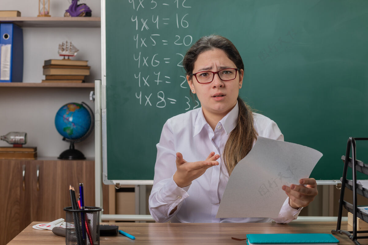 书桌不满意的年轻女老师戴着眼镜坐在课桌旁 拿着白纸检查作业 在教室的黑板前显得很不高兴前面不满意检查