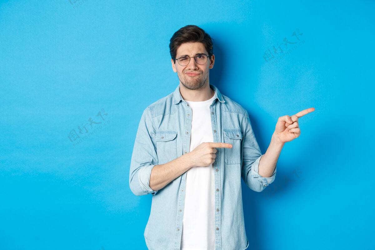 成人戴眼镜的失望的年轻人用手指着版面 展示促销横幅 微笑着表示不满 评判不良产品 站在蓝色背景上男士男性男士