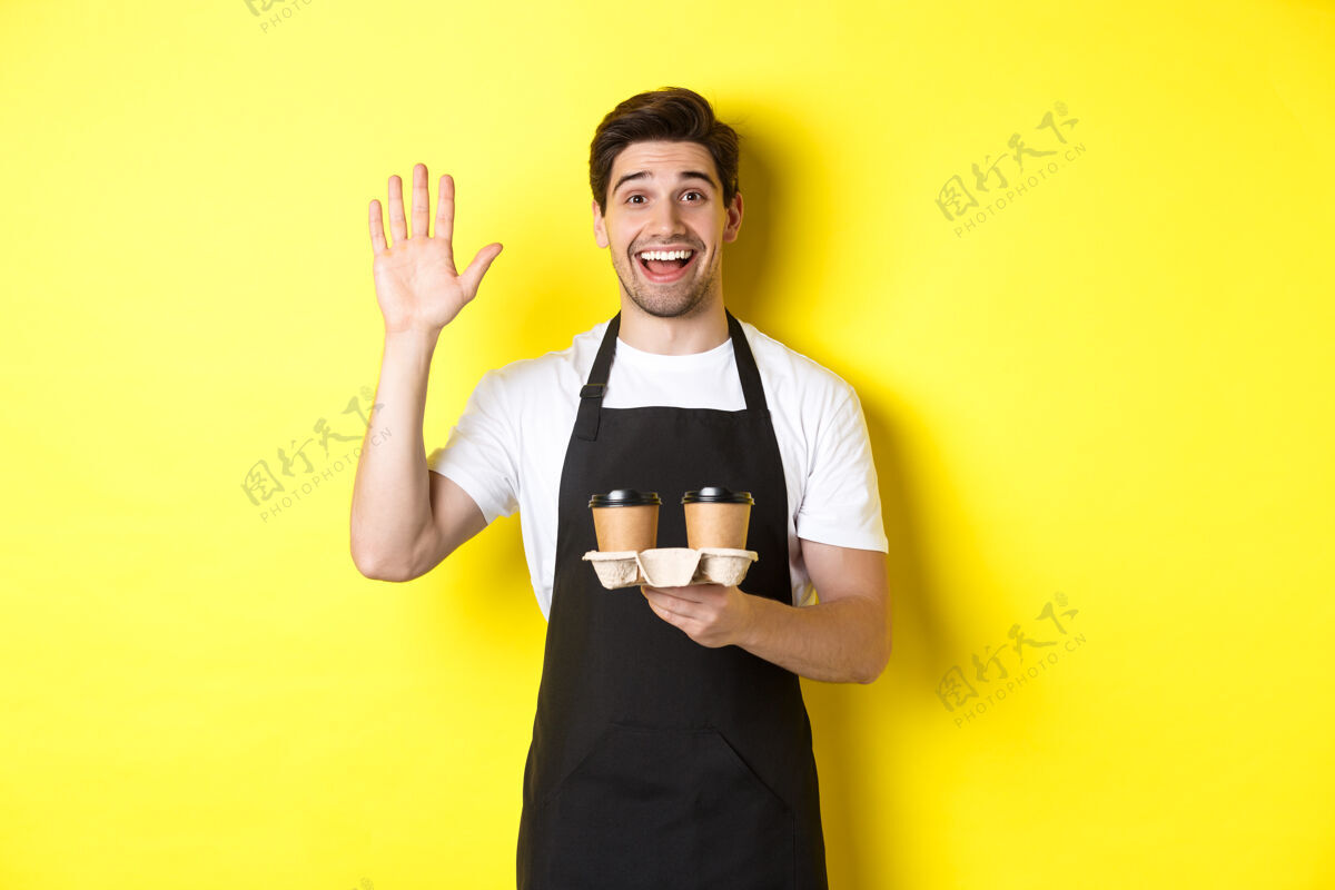 咖啡咖啡馆里友好的服务员向顾客挥手 手里拿着外卖咖啡订单 站在黄色背景下 围着黑色围裙外卖咖啡师时尚