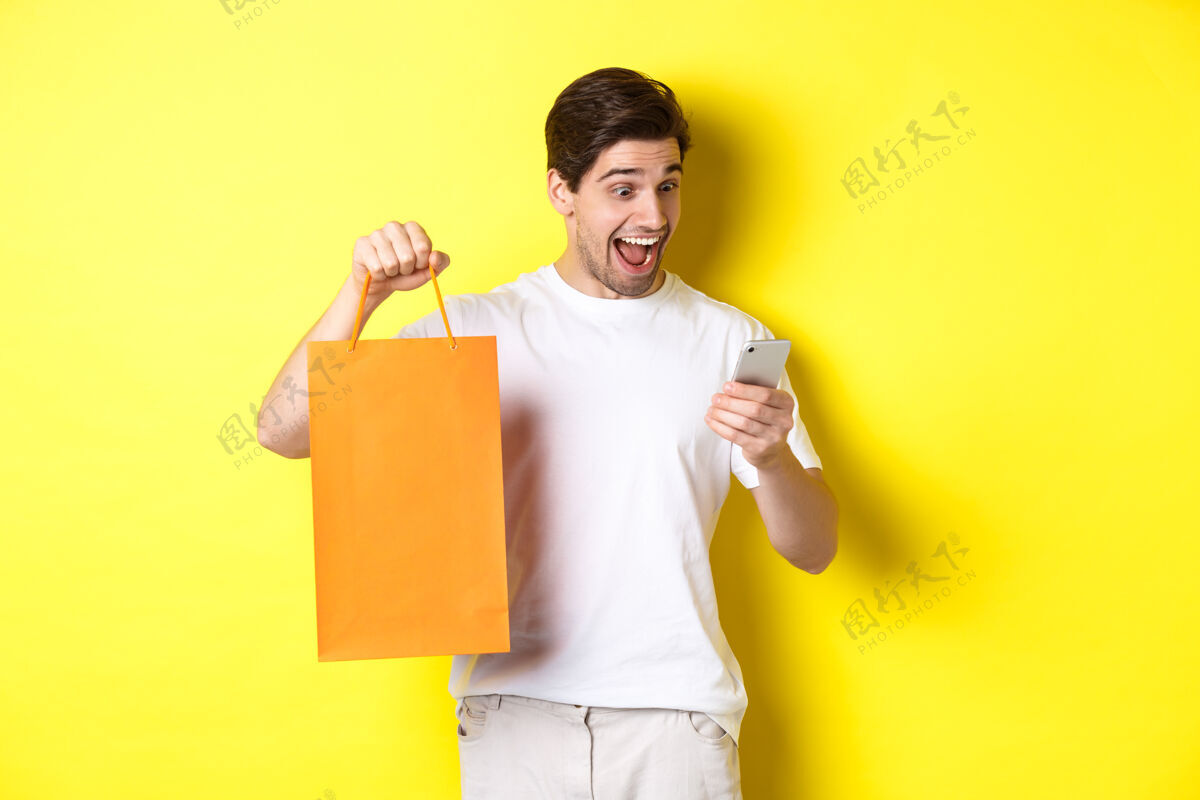 购物袋折扣 网上银行和现金返还的概念惊讶的男人展示购物袋 看着手机屏幕 站在黄色背景下高兴站着表情工作室