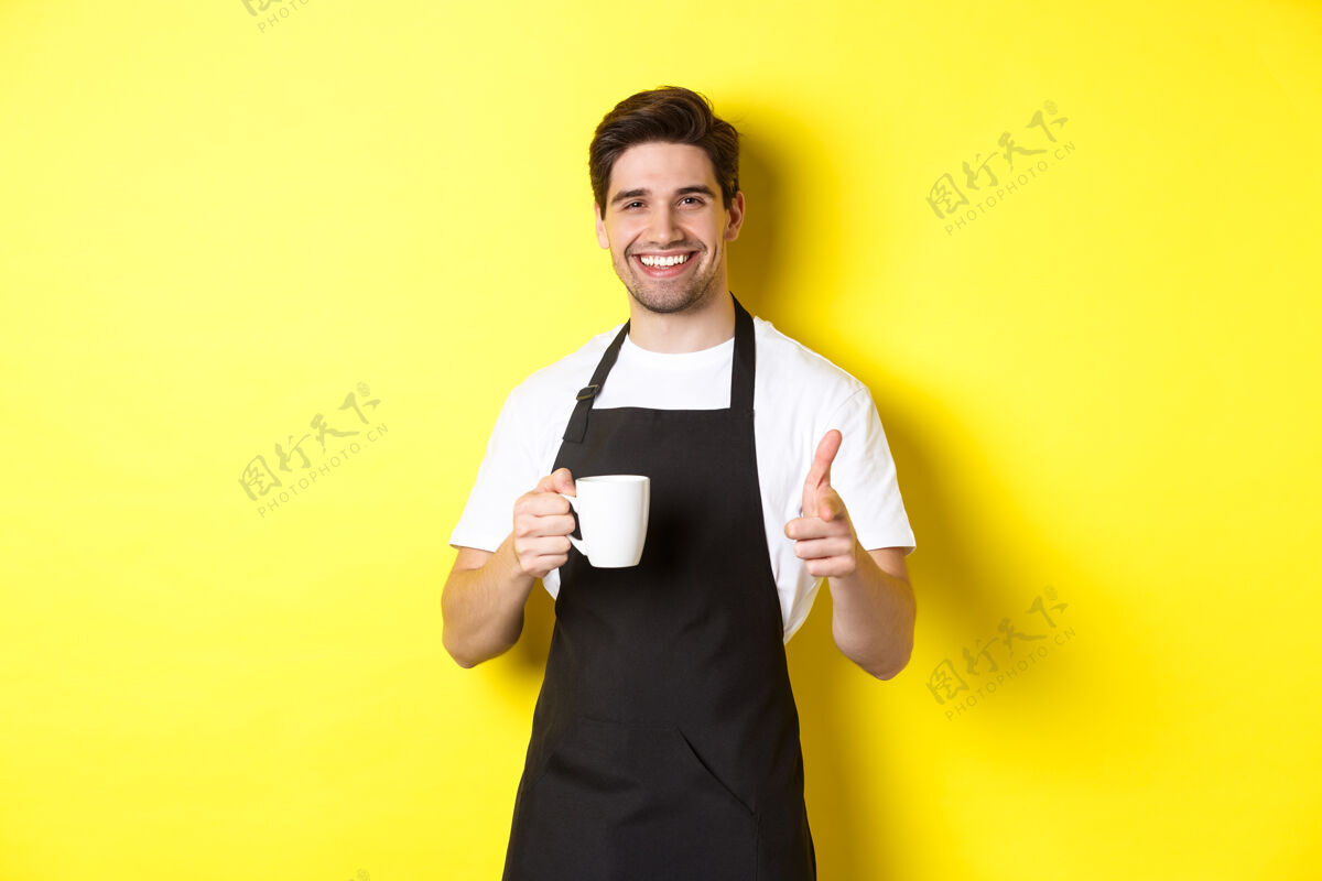 男性咖啡师端着咖啡 用手指枪指着镜头 站在黄色背景下的黑色围裙里男孩黄色时尚