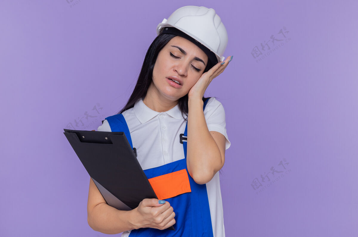 建筑工人身穿施工制服 戴着安全帽 拿着剪贴板的年轻建筑工人站在紫色的墙上 看上去很疲惫 工作过度年轻剪贴板制服