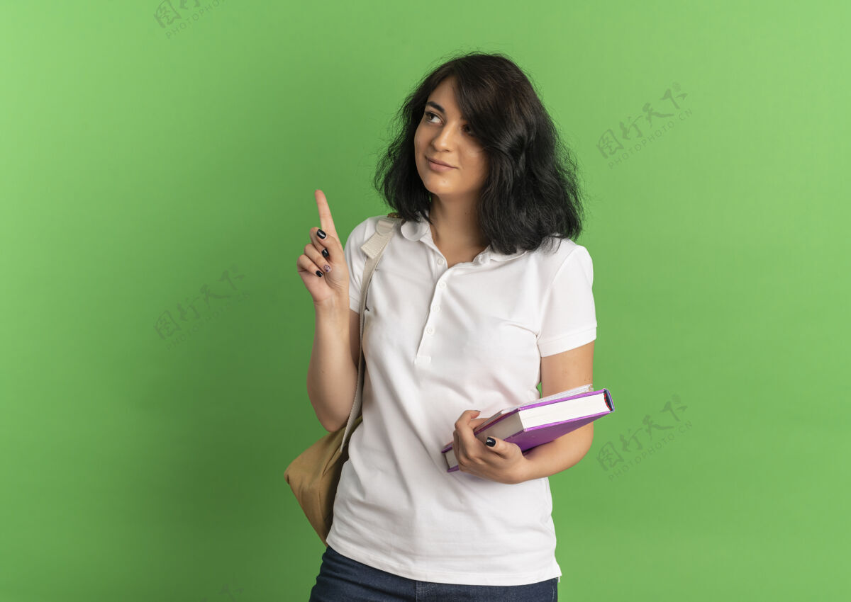拿着年轻漂亮的高加索女学生戴着书包高兴地看了看 指着手里拿着的书 上面有绿色的复印空间绿色向上复制