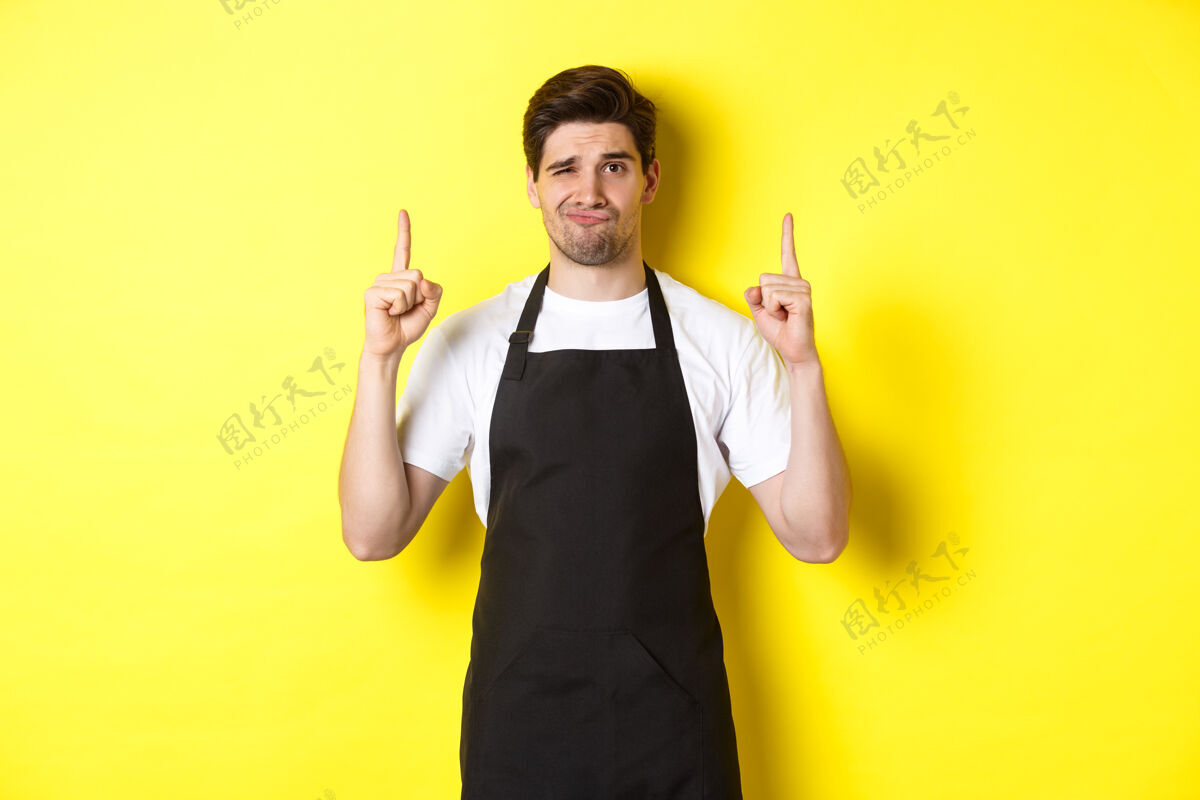 男人不高兴和怀疑的服务员在黑色围裙指着手指起来 表示不喜欢和漠不关心 站在黄色的背景黄色时尚指点