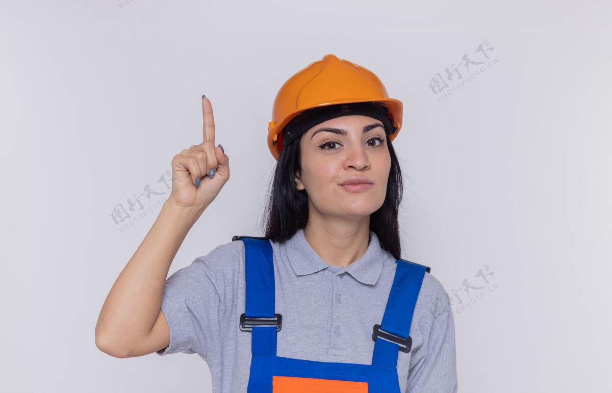 建设者身穿建筑制服 头戴安全帽的年轻建筑工人面带微笑地看着前面 食指站在白色的墙上微笑建筑安全