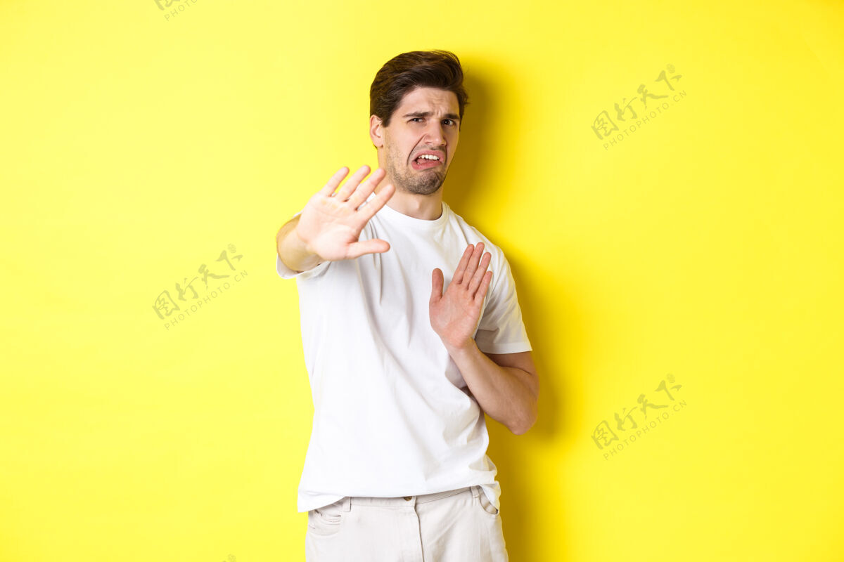 黄色厌恶的男人拒绝 厌恶和厌恶的鬼脸 乞求停止 站在黄色背景下的白色t恤自信工作室厌恶