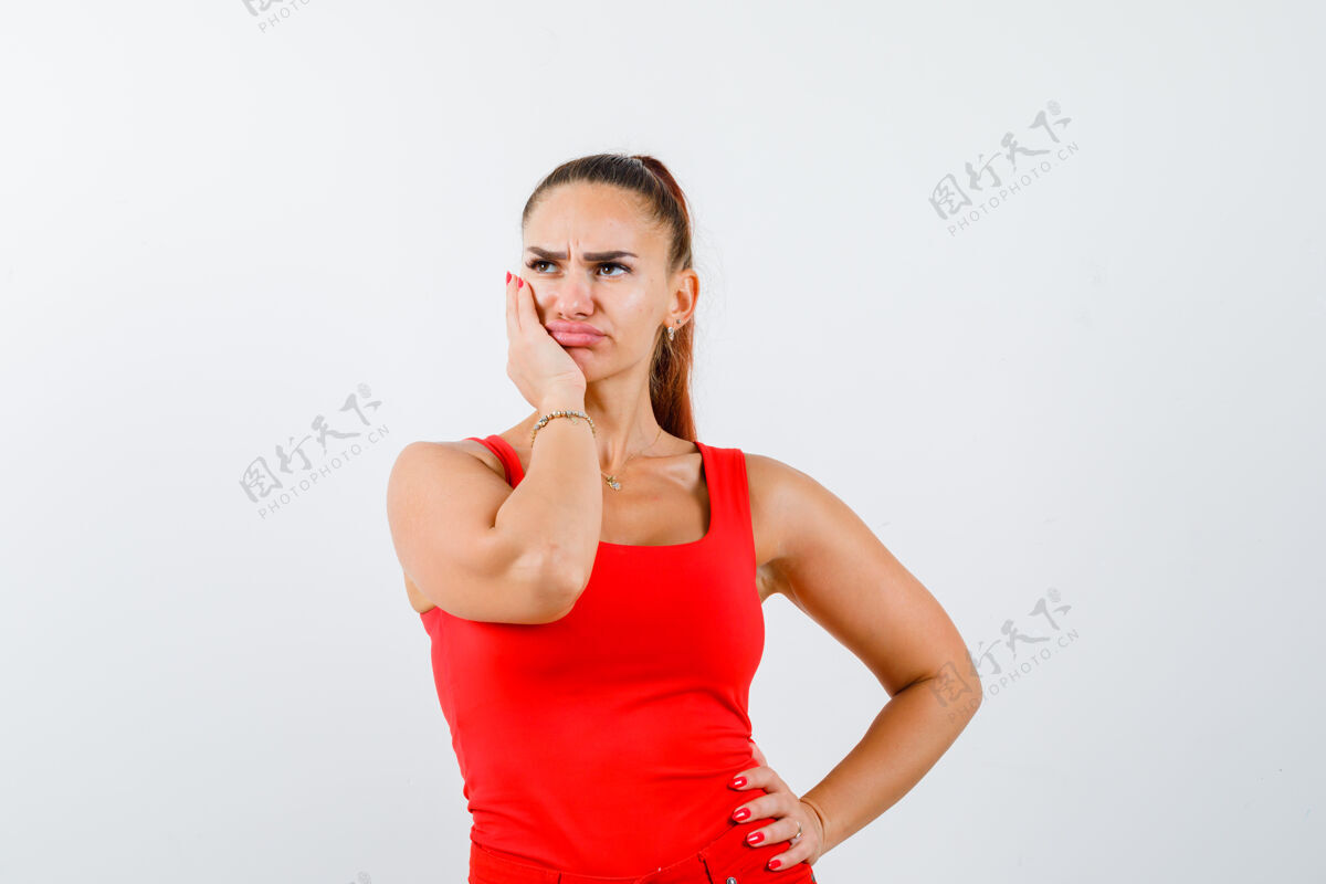 女性年轻的女性手放在脸颊上 手放在腰上 穿着红色的背心 看起来很体贴 前视图脸颊思想护理