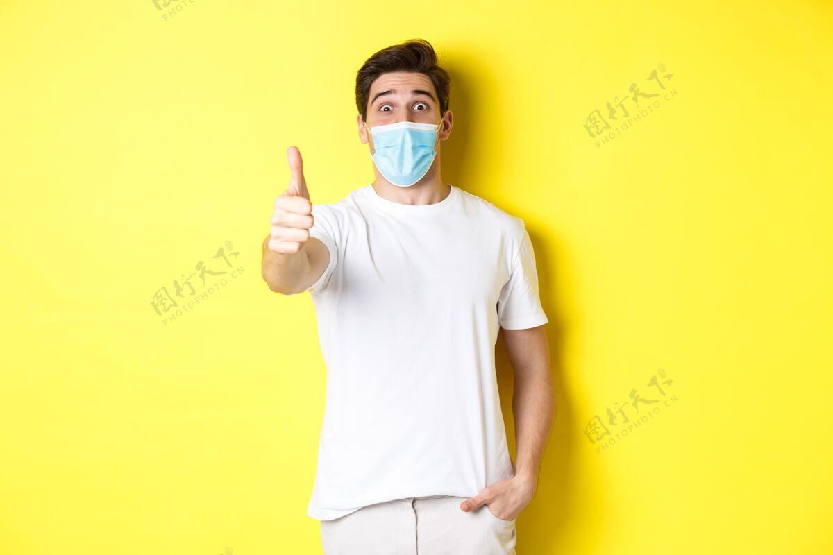 自信冠状病毒 流行病和社会距离的概念令人印象深刻的家伙在医学面具显示拇指赞成 像一些可怕的 黄色的背景面膜成人表情