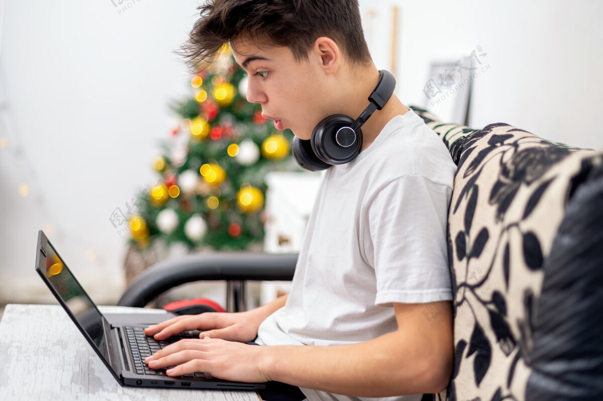 笔记本电脑十几岁的男孩正在家里用带耳机的笔记本电脑圣诞树挂在墙上惊讶的脸长度室内巴德