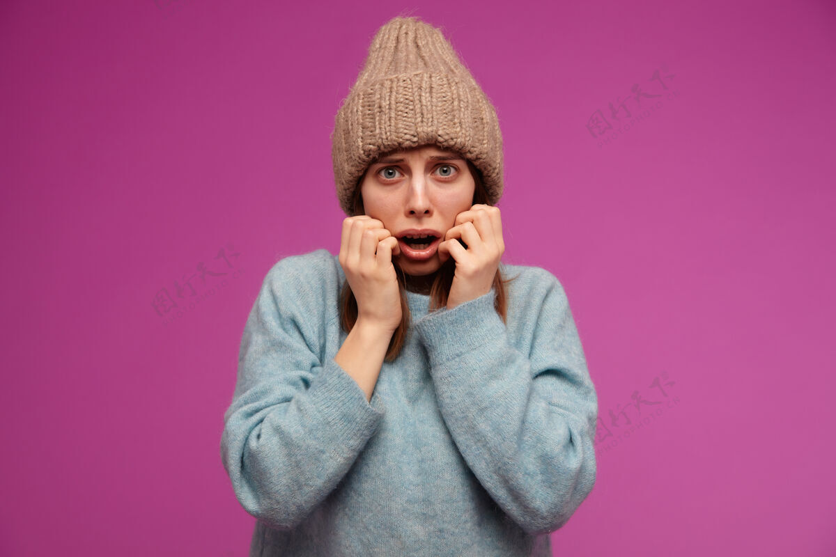 女士一个看起来很害怕的女人 一个被吓坏了的黑发女孩 穿着蓝色的毛衣 戴着编织的帽子 在紫色的墙上恐惧地摸着她的脸自然脸面部