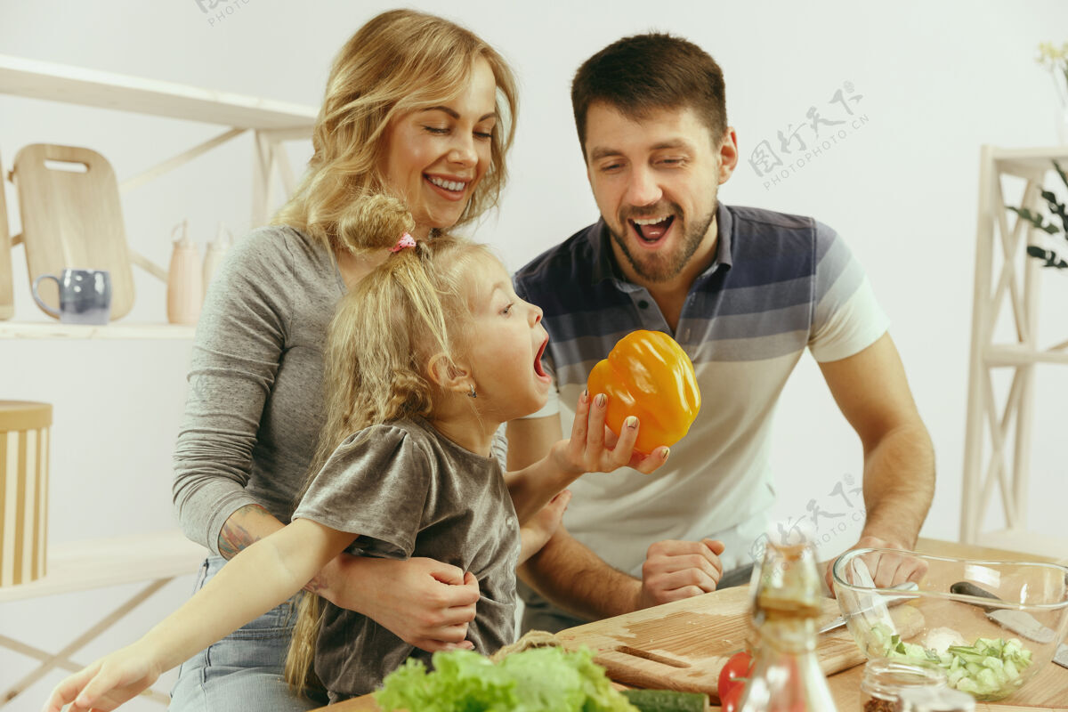 爸爸可爱的小女孩和她美丽的父母在家的厨房里切蔬菜 一边微笑着做沙拉乐趣爱女儿